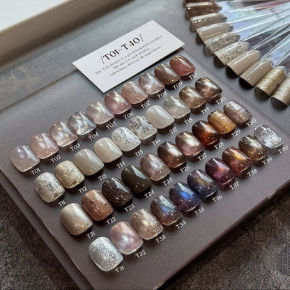 A nail color display tray showing multiple faux nail swatches in various shades and finishes, including neutral beige, pink, brown, gray, and metallic glitter. A small card labeled T01-T04 is placed on top, with additional swatch sticks arranged to the right.